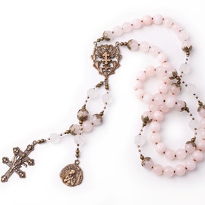 This Saint Joan of Arc rosary is shown on a white background.  Its made with 8mm pink rose quartz and carved clear quartz Our Father beads.  The medals are bronze vintage reproductions.  This design has a side medal of Saint Joan of Arc as well.