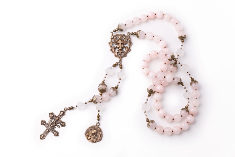 This Saint Joan of Arc rosary is shown on a white background.  Its made with 8mm pink rose quartz and carved clear quartz Our Father beads.  The medals are bronze vintage reproductions.  This design has a side medal of Saint Joan of Arc as well.