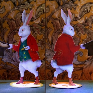 May include: Two white rabbit figurines wearing red and green jackets, holding top hats. The rabbits are standing on circular bases.