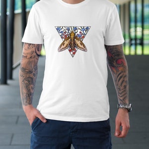 Puede incluir: Una camiseta blanca de manga corta con un dise&ntilde;o gr&aacute;fico central de una polilla marr&oacute;n situada dentro de un tri&aacute;ngulo invertido. La ilustraci&oacute;n incluye dibujos de setas azules y rojas que rodean a la polilla dentro del marco triangular.