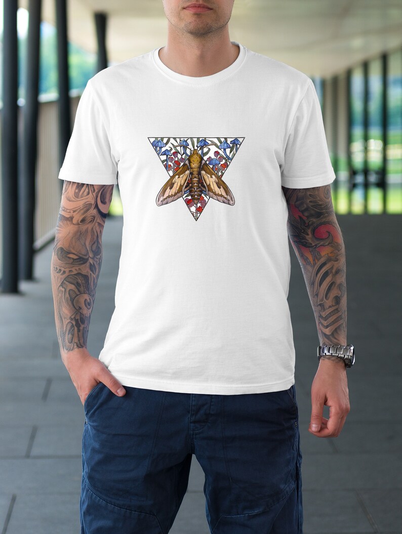Puede incluir: Una camiseta blanca de manga corta con un dise&ntilde;o gr&aacute;fico central de una polilla marr&oacute;n situada dentro de un tri&aacute;ngulo invertido. La ilustraci&oacute;n incluye dibujos de setas azules y rojas que rodean a la polilla dentro del marco triangular.