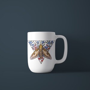 Puede incluir: Una taza de cer&aacute;mica blanca con un dise&ntilde;o gr&aacute;fico triangular. La ilustraci&oacute;n muestra una polilla marr&oacute;n centrada dentro de un tri&aacute;ngulo, rodeada de peque&ntilde;os hongos azules y rojos y follaje verde. La taza tiene un asa curva a la derecha.