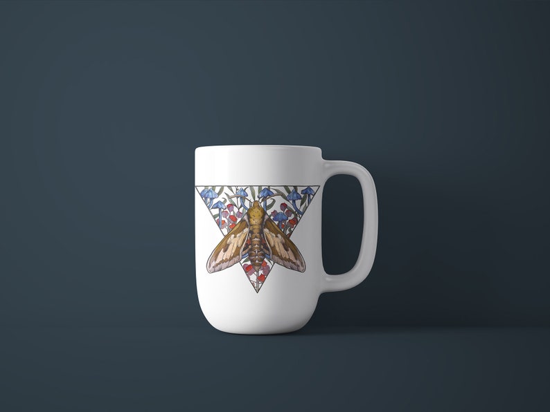 Puede incluir: Una taza de cer&aacute;mica blanca con un dise&ntilde;o gr&aacute;fico triangular. La ilustraci&oacute;n muestra una polilla marr&oacute;n centrada dentro de un tri&aacute;ngulo, rodeada de peque&ntilde;os hongos azules y rojos y follaje verde. La taza tiene un asa curva a la derecha.
