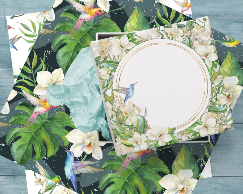 May include: A white gift box with a gold circle frame and a watercolor design of white orchids, green leaves, and blue and orange hummingbirds. The box is open and the inside is white.