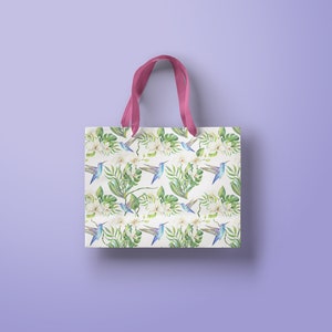 May include: A white tote bag with pink handles features a repeating pattern of blue hummingbirds, white flowers, and green leaves.