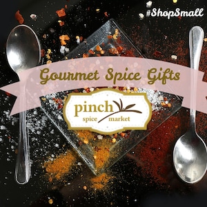 May include: Gourmet spice gifts from Pinch Spice Market. A close-up of a glass plate with various spices, including red pepper flakes, salt, and turmeric. The plate is surrounded by scattered spices on a black background. Two silver spoons are on either side of the plate.