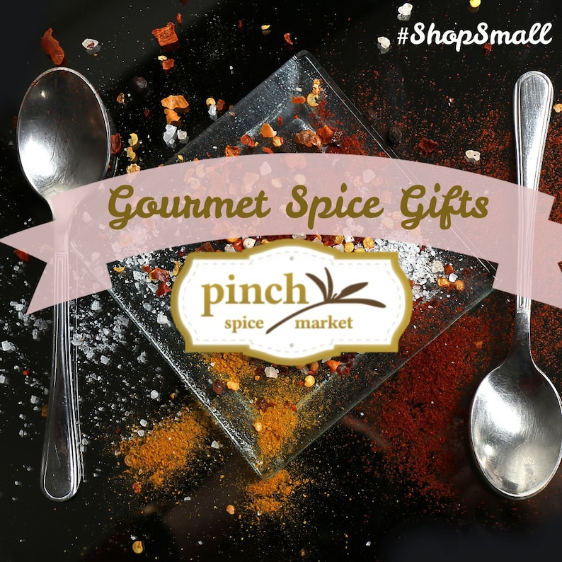 May include: Gourmet spice gifts from Pinch Spice Market. A close-up of a glass plate with various spices, including red pepper flakes, salt, and turmeric. The plate is surrounded by scattered spices on a black background. Two silver spoons are on either side of the plate.