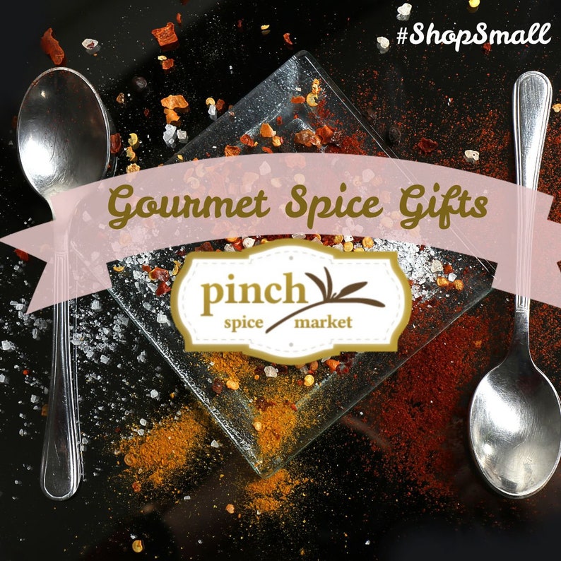 May include: Gourmet spice gifts from Pinch Spice Market. A close-up of a glass plate with various spices, including red pepper flakes, salt, and turmeric. The plate is surrounded by scattered spices on a black background. Two silver spoons are on either side of the plate.