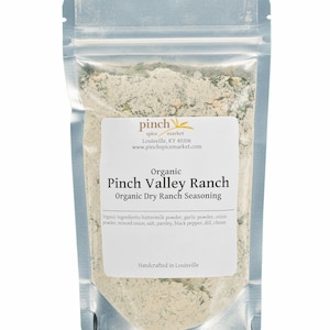 May include: A silver resealable pouch filled with a light brown and green organic dry ranch seasoning mix. The pouch has a white label with the text "pinch spice market" and "Organic Pinch Valley Ranch Organic Dry Ranch Seasoning". The label also lists the ingredients and states "Handcrafted in Louisville".
