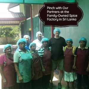 May include: A group of people wearing hairnets and aprons standing in front of a building. The text overlay reads: Pinch with Our Partners at the Family-Owned Spice Factory in Sri Lanka. The individuals are dressed in various shirts and aprons.