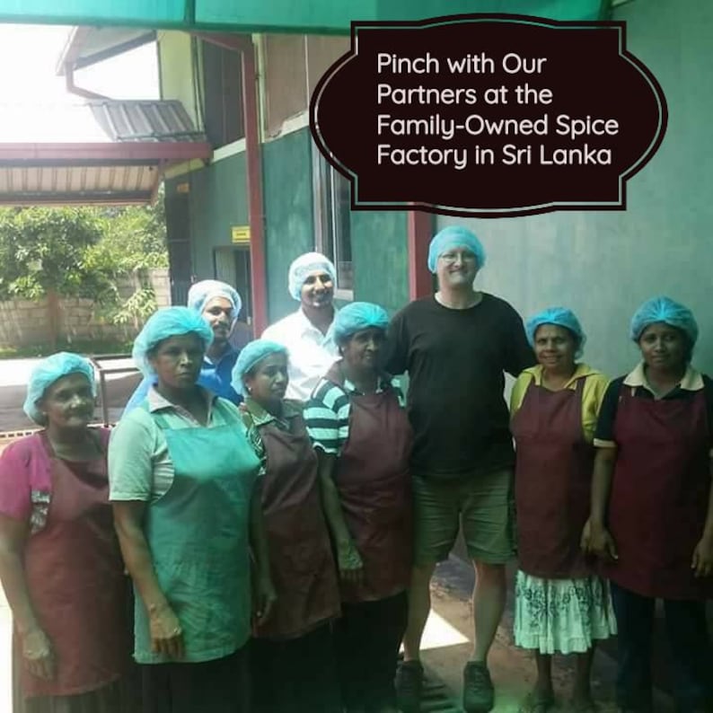 May include: A group of people wearing hairnets and aprons standing in front of a building. The text overlay reads: Pinch with Our Partners at the Family-Owned Spice Factory in Sri Lanka. The individuals are dressed in various shirts and aprons.