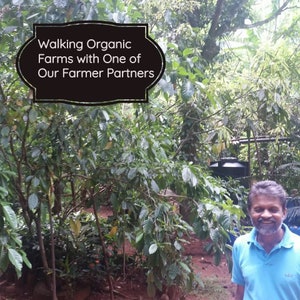 May include: A person with a beard wearing a light blue polo shirt stands in a lush green organic farm. A black sign with a decorative border features the white text: Walking Organic Farms with One of Our Farmer Partners, set against trees.