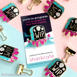 May include: A set of five enamel pins featuring a tombstone with the text "WIP" (Work in Progress) in gold lettering, dripping pink and blue paint. The pins are designed with a skull and crescent moon on the tombstone. The text "work-in-progress" is written on a black card with white text, along with the text "a pin for people who never finish anything". The card also includes the text "pretty pins for petty people by sharkcafe" and the website address "sharkcafeart.com".