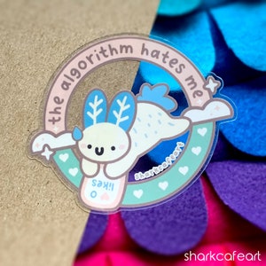 May include: A sticker featuring a cartoon axolotl with blue fins and a pink heart. The axolotl is holding a pink and white sign that says "Say!!" The axolotl is surrounded by a circle with the text "the algorithm hates me" in pink and green.