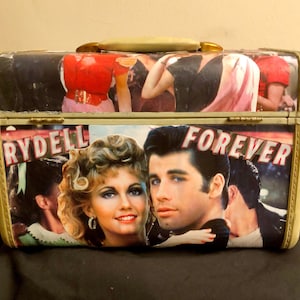 May include: A vintage-style, cream-colored suitcase with a black and white image of a couple on the front. The suitcase has a gold handle and gold accents. The image on the front of the suitcase includes the words "Rydell" and "Forever".