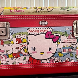 May include: A red, rectangular, hard-sided, Hello Kitty-themed makeup case with a silver lock and clasp. The case has a colorful print of Hello Kitty characters and the words "candy cand" and "Hook-A-Duck".