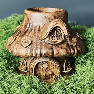 Op de afbeelding: Een bruin keramisch paddenstoelhuis met een textuuroppervlak, hartvormige accenten en gewelfde ramen en deuren. Het huis staat op een bedje van groen mos, wat een grillige, sprookjesachtige esthetiek creëert.