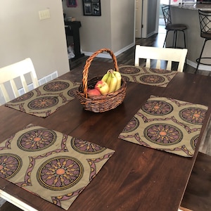 May include: A dark wooden dining table set with four patterned placemats and a woven basket filled with fruit. The placemats have a circular design in shades of olive green, purple, and gold. The basket contains bananas and apples.