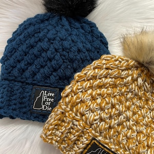 May include: Two crocheted beanies with pom-poms. One is dark blue with a black pom-pom, and the other is a mustard yellow and cream blend with a tan pom-pom. Each features a black rectangular patch with the text Live Free or Die and a state outline.