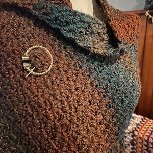 Può includere: Uno scialle a collo alto lavorato all'uncinetto a mano, realizzato con un filato variegato nei toni del marrone scuro, rame e ottanio. Il capo è fissato con una spilla circolare in metallo dorato con una texture intrecciata e chiusura a spillo.