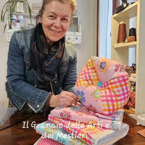 Può includere: Una donna sorride, in piedi dietro una pila di tessuti colorati e un cuscino a fantasia floreale. I tessuti includono motivi rosa, arancioni e blu. Il testo "Il Granaio delle Arti e dei Mestieri" è visibile.