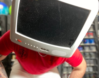 May include: A person wearing a red shirt with a black and white logo and white pants. The person is wearing a white computer monitor on their head. The monitor has a black screen.