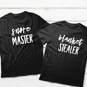 Puede incluir: Dos camisetas negras con texto blanco. Una camiseta dice "snore master" y la otra dice "blanket stealer".
