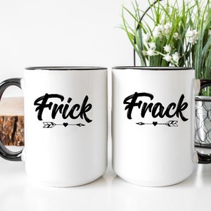 May include: Two white ceramic mugs with black rims and handles. The mug on the left says "Frick" with an arrow and heart. The mug on the right says "Frack" with an arrow and heart.
