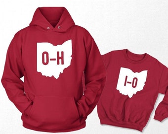May include: Two red sweatshirts, a hooded top and a crew neck, each displaying a white outline of the state of Ohio. The hooded top has the letters "O-H" inside the state outline, and the crew neck has "1-O".