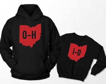 Pode incluir: Duas camisolas pretas, uma com capuz e outra de gola redonda, ambas com um contorno vermelho de Ohio. A camisola com capuz exibe "O-H" e a camisola de gola redonda "1-0" em preto. Vestuário.