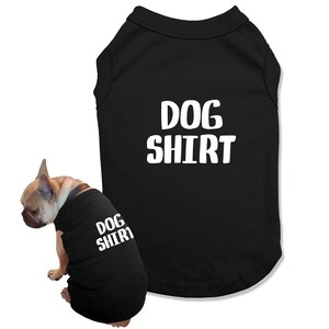 May include: Black dog shirt with the text "DOG SHIRT" in white. A small dog is wearing a matching shirt. The dog is light brown with a black collar.
