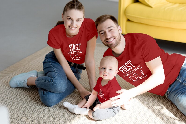 May include: A family of three wearing matching red shirts with white text. The shirts say "First Christmas as Mom", "First Christmas as Dad", and "First Christmas".