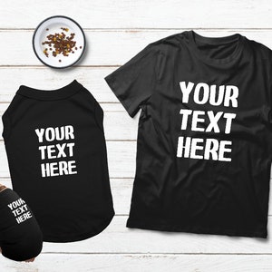 May include: Black t-shirts and dog shirts with the text "YOUR TEXT HERE" in white. A small dog is wearing a matching shirt. A bowl of dog food is in the background.