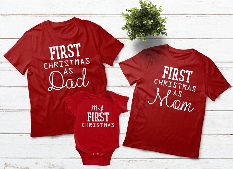 May include: Red Christmas shirts for a family of three. The adult shirts say "First Christmas as Dad" and "First Christmas as Mom". The baby onesie says "My First Christmas".