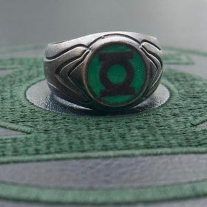 May include: A silver-toned ring featuring the Green Lantern symbol. The ring has a detailed design with a green and black emblem at the center. The ring is set against a green and gray background.