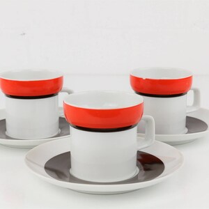 Könnte beinhalten: Drei weiße Keramik-Kaffeetassen mit roten und schwarzen Bändern. Jede Tasse steht auf einer passenden Untertasse mit einem schwarz-weißen Design.