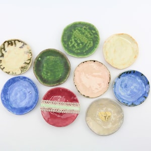 Könnte beinhalten: Neun kleine Keramikteller in verschiedenen Farben, darunter Blau, Grün, Rot, Rosa und Gelb. Die Teller sind alle in Form und Größe leicht unterschiedlich und einige haben dekorative Details.