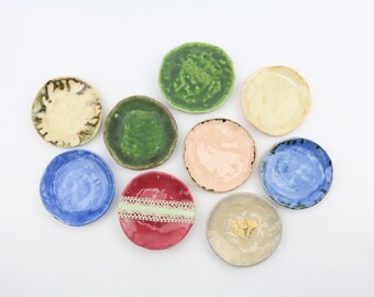Op de afbeelding: Negen kleine keramische schoteltjes in verschillende kleuren, waaronder blauw, groen, rood, roze en geel. De schoteltjes zijn allemaal iets anders van vorm en grootte, en sommige hebben decoratieve details.