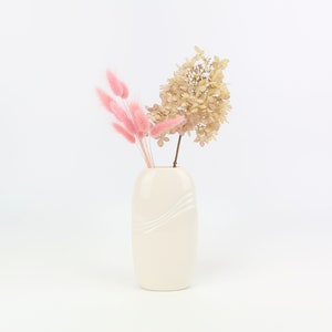 Könnte beinhalten: Eine weiße Keramikvase mit wellenförmigem Design enthält einen Strauß getrockneter Blumen. Die Blumen sind eine Mischung aus rosa und beige.