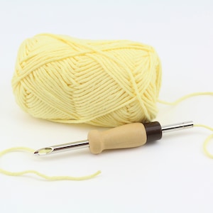 Pode incluir: Uma bola de fio amarelo claro e uma agulha de lã com cabo de madeira e um gancho de metal.