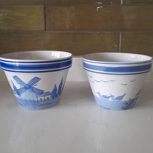 May include: Two white ceramic flower pots with blue and white designs. One pot features a windmill and the other features birds.