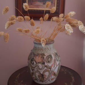 May include: A ceramic vase with a floral pattern, holding dried wheat, is displayed on a small, round table. The table is dark brown with painted floral details. A framed painting is visible in the background.