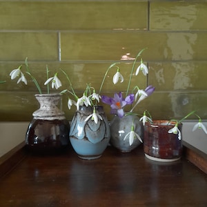 以下が含まれることがあります： 木製の表面に、さまざまな色とスタイルの小さなセラミックの花瓶が5つ並んでいます。それぞれの瓶には、白いスノードロップの花と紫色のクロッカスの花束が活けてあります。