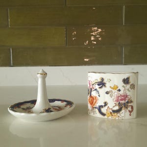 May include: A white ceramic ring holder with a gold-tipped central post and a decorative dish base. Beside it is a matching floral-patterned ceramic container with gold trim. The items are on a white surface.