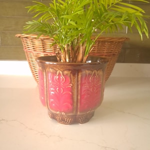 May include: A ceramic plant pot featuring a vibrant hot pink centre with embossed vertical leaf patterns, framed by a dark brown glazed rim and base. A green palm plant grows from the pot, which sits in front of a woven brown wicker basket.