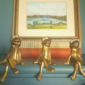 May include: Three brass frog ornaments with outstretched legs, sitting on a shelf. The frogs have a smiling expression and are positioned in front of a framed landscape painting and a stack of books.