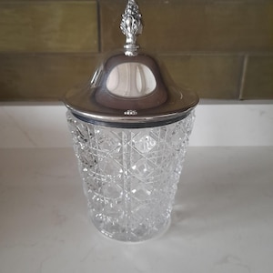 May include: A clear glass jar with a silver lid and a decorative finial. The jar has a diamond pattern.