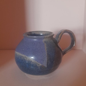 May include: A handmade ceramic mug with a rounded body and a handle. The mug features a blue and purple colour scheme, with a speckled texture. The handle is a darker shade of blue. The mug is likely used for drinking beverages.