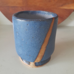 May include: A blue ceramic tumbler with a tan stripe. The small cup has a flared base and a speckled texture. The interior is a darker blue. The tumbler is suitable for hot or cold drinks.