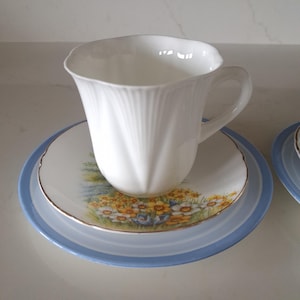 May include: A white teacup with a fluted design sits on a saucer with a floral pattern and a light blue rim. The teacup has a curved handle. The saucer is placed on a larger light blue plate.